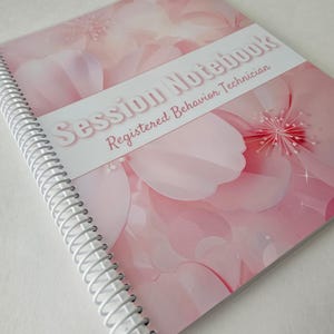 May include: A spiral-bound notebook with a soft pink floral background. The cover features the white text Session Notebook and the smaller pink text Registered Behavior Technician. The design includes layered petal graphics and subtle star accents.