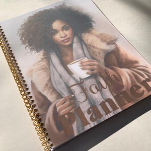 May include: A spiral bound planner with a brown and white illustration of a person holding a cup of coffee. The text "Faith Planner" is printed in gold on the cover.