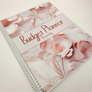 May include: A spiral-bound budget planner with a white and rose gold marble background featuring large, soft pink floral illustrations. The cover displays the text Budget Planner Bi-weekly in a dark rose gold script font across the center.