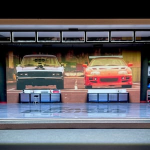 May include: A model garage diorama with a black and white checkered floor, featuring a black Dodge Charger and a red sports car on the back wall. The garage has a black metal door and two sets of black metal shelves on either side of the cars.