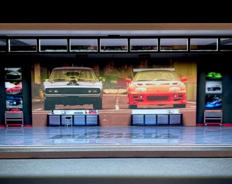 May include: A model garage diorama with a black and white checkered floor, featuring a black Dodge Charger and a red sports car on the back wall. The garage has a black metal door and two sets of black metal shelves on either side of the cars.