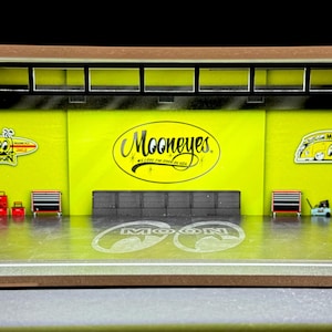 May include: A yellow and black diorama of a garage with a Mooneyes logo, a yellow car, and a yellow and black hot rod. The garage has a black metal frame and a wood floor.