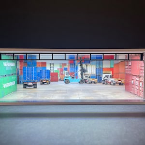 May include: A model of a warehouse with a large open doorway revealing a scene of a shipping container yard with a forklift and several trucks. The containers are stacked high and are painted in various colors, including red, blue, and green. The containers have the word "EVERGREEN" printed on them.