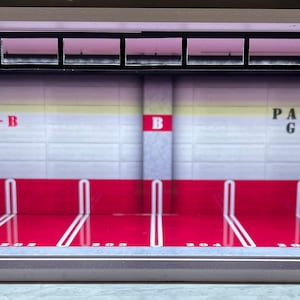 Op de afbeelding: Een witte en rode modelgarage met een zwart metalen frame en een bord met de tekst "Parking Garage". De garage heeft een witte betegelde muur met een rode streep aan de onderkant en een gele streep aan de bovenkant. De garage heeft twee parkeerplaatsen met witte lijnen op de vloer.