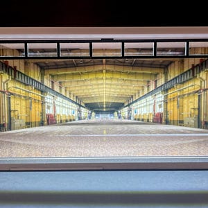 May include: A miniature diorama depicting the interior of an industrial warehouse. The model shows a long, open space with yellow walls, a concrete floor, and a high ceiling with visible support beams. The model has a white roof and a black window frame.