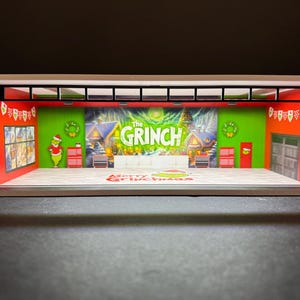 May include: A miniature stage set with a green and red backdrop featuring the words "The Grinch" and a festive scene. The stage is decorated with Christmas lights and a sign that reads "Merry Grinchmas".
