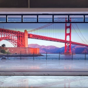 Könnte beinhalten: Ein Miniatur-Diorama mit einer Kulisse der Golden Gate Bridge in San Francisco. Das Diorama besteht aus Holz und Glas und hat eine weiße Basis. Die Brücke ist in Rot und Orange bemalt und vor einem blauen Himmel mit weißen Wolken platziert.