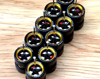 May include: A collection of miniature black tires with gold rims and silver pins. The tires have a red accent and white lettering. The tires are stacked on top of each other on a wooden surface.