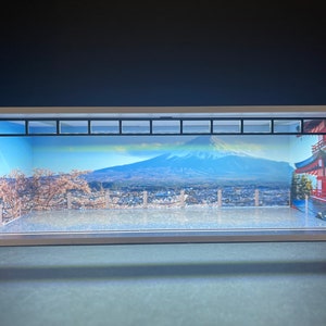 Pode incluir: Um modelo branco de um edifício com uma fachada de vidro, mostrando um cenário cênico do Monte Fuji, uma cidade e flores de cerejeira. O modelo é iluminado por dentro, criando um brilho quente.