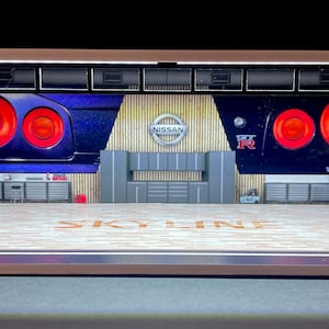 Puede incluir: Un diorama de garaje modelo con suelo y paredes de madera. Las paredes están decoradas con imágenes de coches Nissan Skyline R34. El diorama tiene un marco de metal negro y una parte delantera de plástico transparente.