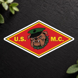 May include: A diamond-shaped sticker featuring a red background with a yellow border. In the center is a brown bulldog wearing a green military cap and a spiked collar. The text U.S. M.C. is printed in yellow on either side of the bulldog.