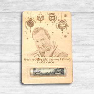 May include: Wooden gift card holder with a humorous engraved image of a man making an 'okay' gesture. The design includes Christmas ornaments and the phrase "Get yourself something real nice..." A rolled-up dollar bill is visible.