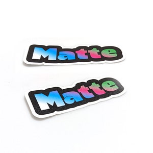 May include: Two stickers with the word "Matte" in a gradient of blue, pink, and green colours. The letters are outlined in black and have a textured, almost glittery appearance.