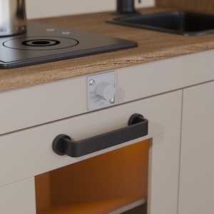 Peut inclure: Une cuisine jouet blanche avec une plaque de cuisson noire, un évier noir et deux portes de placard noires avec des poignées noires. La plaque de cuisson a un bouton argenté et une casserole argentée sur le dessus. Les portes des placards ont un bouton argenté sur la porte supérieure gauche.