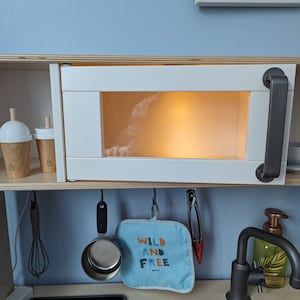 May include: A white toy microwave with a grey handle and a clear window. The microwave is lit up from inside. There are two wooden cups on the shelf above the microwave. The microwave is mounted on a wooden shelf.