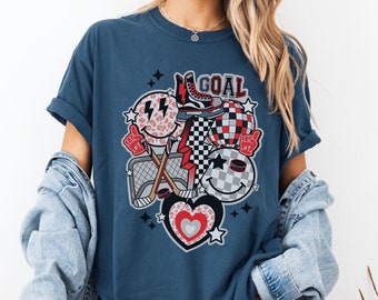 May include: A navy blue t-shirt with a graphic design featuring a hockey stick, a puck, a heart, and smiley faces. The graphic also includes the word "GOAL".