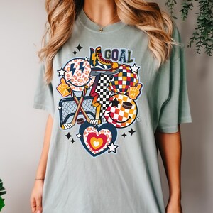 May include: A light blue t-shirt with a graphic design featuring a hockey stick, a puck, a heart, a star, a checkered ball, and a smiley face with a lightning bolt. The text "GOAL" is printed on the shirt.
