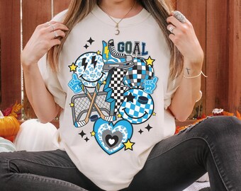 May include: A white t-shirt with a blue, black, and white graphic design featuring a hockey stick, a puck, a heart, and a smiley face. The graphic also includes the word "GOAL" in black text.