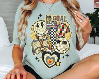 May include: A light green t-shirt featuring a hockey-themed graphic. The design includes a hockey skate, a hockey net, crossed hockey sticks, a checkered pattern, smiley faces, and the word "GOAL".