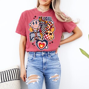 May include: A red t-shirt with a graphic design of hockey equipment, a smiley face, a heart, and the word "GOAL".