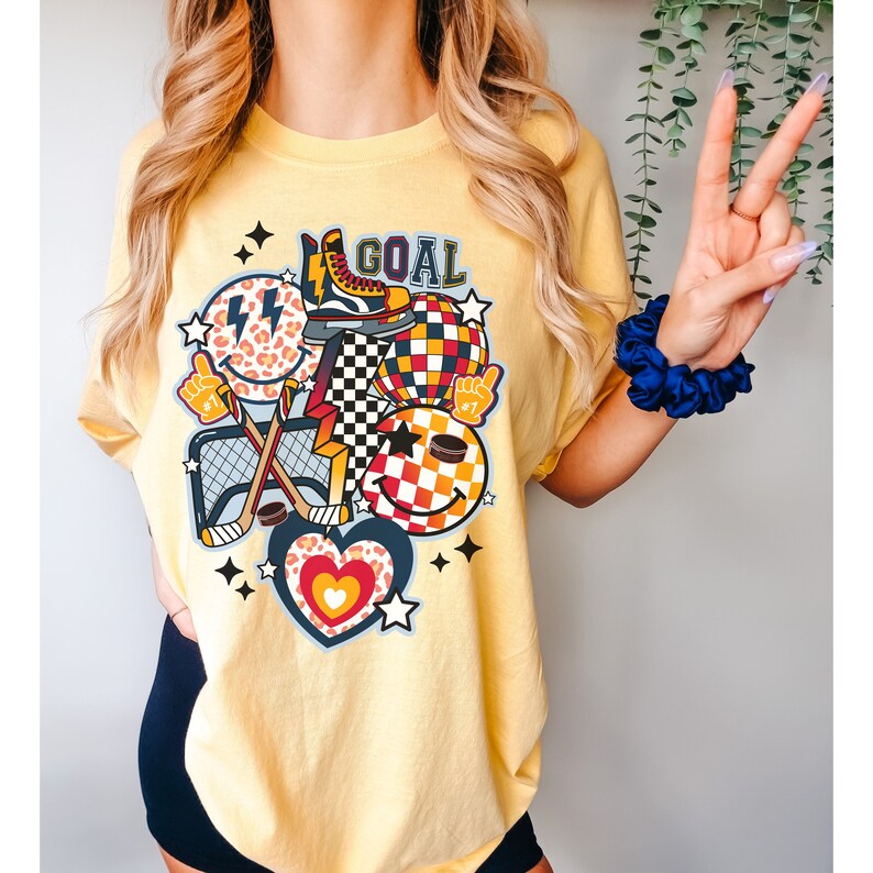 May include: Yellow t-shirt with a graphic design featuring a hockey stick, a net, a puck, a smiley face with a checkered pattern, a heart with a leopard print, and the word "GOAL".