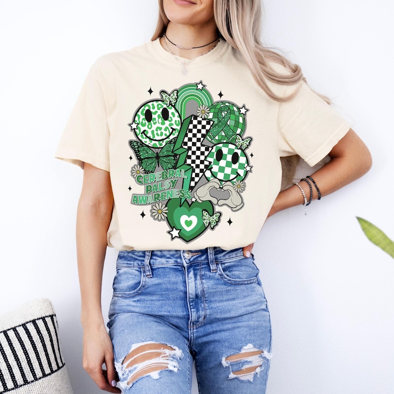 May include: A cream coloured t-shirt with a green, black and white graphic design. The design features a smiley face, a lightning bolt, a chequered pattern, a heart and butterflies. The text "Cerebral Palsy Awareness" is printed on the shirt.