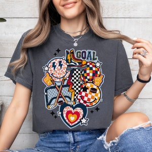May include: A dark gray t-shirt with a graphic design featuring a hockey stick, a net, a puck, a smiley face, a heart, and a star. The text "GOAL" is printed on the shirt.