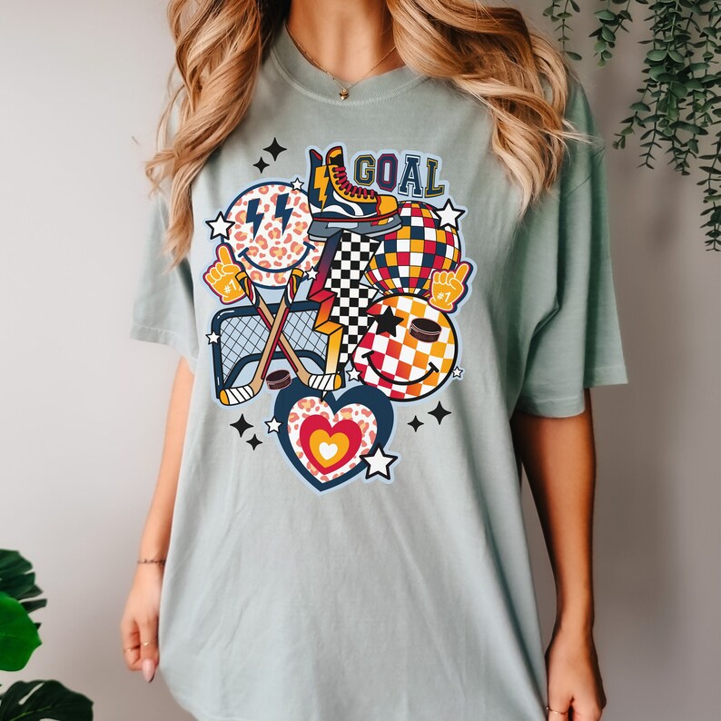May include: A light blue t-shirt with a graphic design featuring a hockey stick, a puck, a heart, a star, a checkered ball, and a smiley face with a lightning bolt. The text "GOAL" is printed on the shirt.