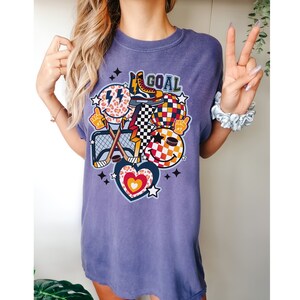 May include: Purple t-shirt with a graphic design featuring a hockey stick, a heart, a smiley face, a checkered pattern, and the word "GOAL".