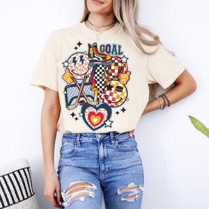 May include: A cream colored t-shirt with a graphic design featuring a hockey stick, a net, a smiley face, a heart, a checkerboard pattern, a star, a lightning bolt, a leopard print, a skate, and the word "GOAL".