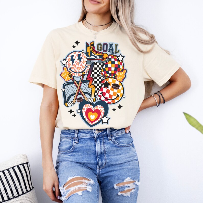 May include: A cream colored t-shirt with a graphic design featuring a hockey stick, a net, a smiley face, a heart, a checkerboard pattern, a star, a lightning bolt, a leopard print, a skate, and the word "GOAL".