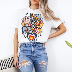 May include: White t-shirt with a colorful graphic design featuring a hockey stick, a puck, a heart, a smiley face, and the word "GOAL".