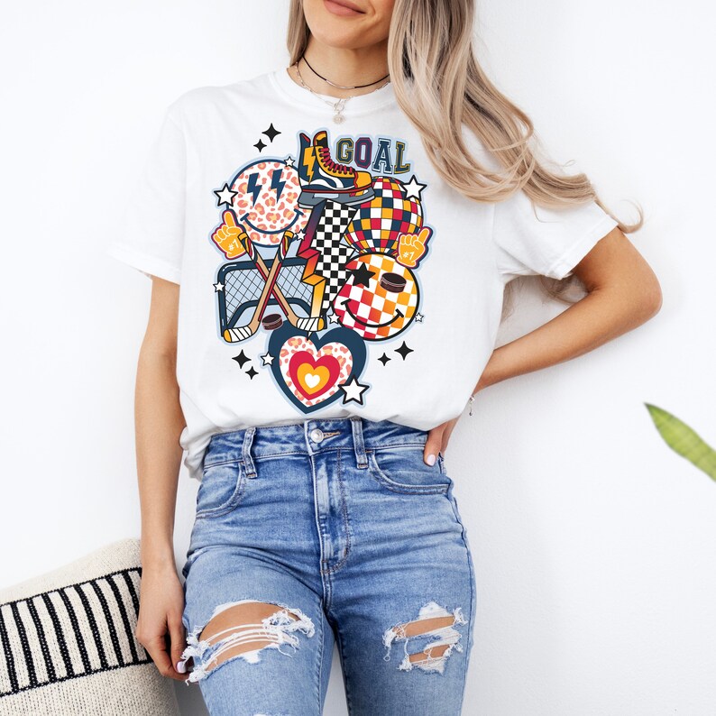 May include: White t-shirt with a colorful graphic design featuring a hockey stick, a puck, a heart, a smiley face, and the word "GOAL".