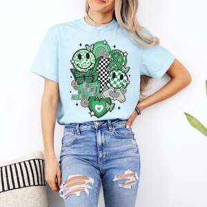May include: A light blue t-shirt with a green, black and white graphic design. The graphic features a variety of smiley faces, butterflies, and the text "Celebrate Cerebral Palsy Awareness".
