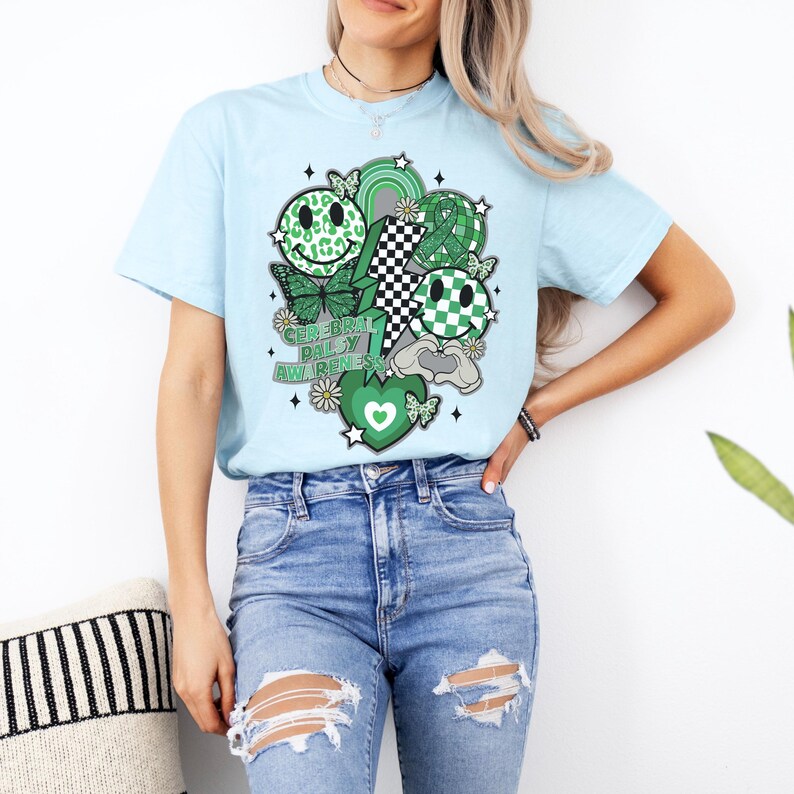 May include: A light blue t-shirt with a green, black and white graphic design. The graphic features a variety of smiley faces, butterflies, and the text "Celebrate Cerebral Palsy Awareness".