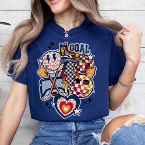 May include: A navy blue t-shirt with a graphic design featuring a hockey stick, a puck, a skate, a heart, and smiley faces with a checkered pattern. The text "GOAL" is printed on the shirt.