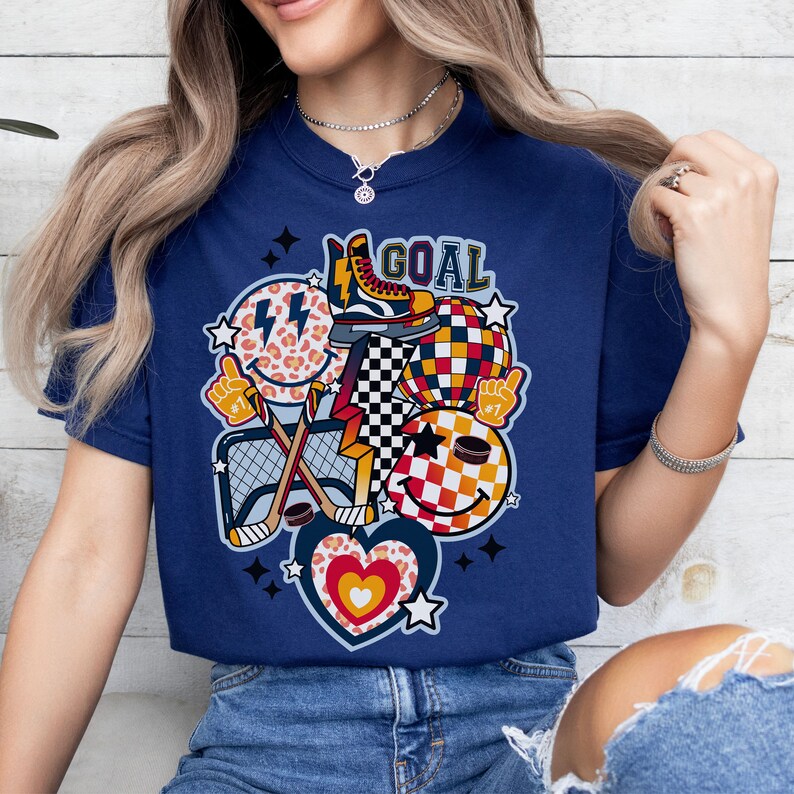 May include: A navy blue t-shirt with a graphic design featuring a hockey stick, a puck, a skate, a heart, and smiley faces with a checkered pattern. The text "GOAL" is printed on the shirt.