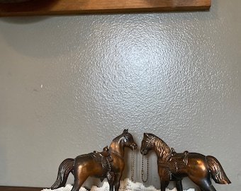 May include: Two bronze-colored horse figurines with saddles and bridles. The horses are connected by a chain. The figurines are on a white lace tablecloth. The background includes a wooden cutting board and a gray wall.