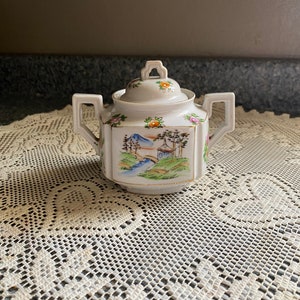 May include: A white porcelain sugar bowl with a lid, depicting a landscape scene with a bridge, river, and mountains. The bowl has two handles and is decorated with floral accents in orange, pink, and green. The rim is trimmed with gold.