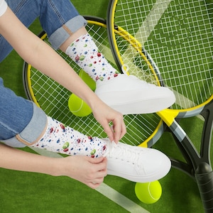 Può includere: Una persona che indossa scarpe da ginnastica bianche con calzini con schizzi di vernice colorata, seduta su un campo da tennis con due racchette da tennis e due palline da tennis.
