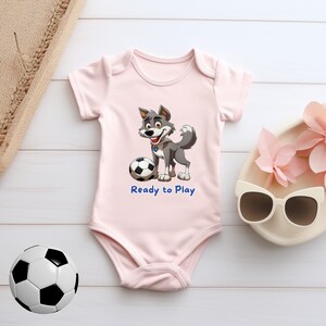 May include: A pink baby onesie with a cartoon gray wolf wearing a blue collar and holding a soccer ball. The onesie has the text "Ready to Play" printed in blue.