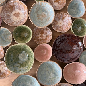 May include: A selection of handcrafted ceramic bowls in assorted sizes and hues. The bowls display a range of earthy tones, such as brown, green, blue, and pink. The bowls have uneven edges and a rustic aesthetic, indicating a handmade origin.