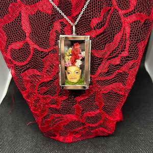 Pode incluir: Um colar com um pingente de prata, com uma frente de vidro que apresenta um pequeno rosto pintado com flores vermelhas dentro. O pingente está sobre um fundo de renda vermelha.