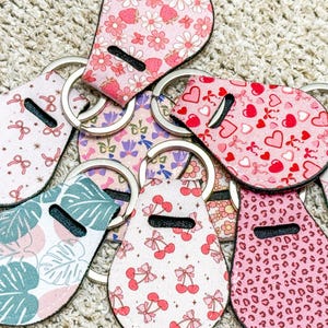 K&ouml;nnte beinhalten: Eine Sammlung von acht rosa und wei&szlig;en Schl&uuml;sselanh&auml;ngern mit verschiedenen Mustern, darunter Herzen, Schleifen, Blumen und Kirschen. Die Schl&uuml;sselanh&auml;nger sind aus Stoff und haben silberne Schl&uuml;sselringe.