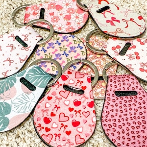 K&ouml;nnte beinhalten: Eine Sammlung bunter Schl&uuml;sselanh&auml;nger mit verschiedenen Mustern, darunter Herzen, Blumen und geometrische Designs. Die Schl&uuml;sselanh&auml;nger sind aus Stoff gefertigt und haben einen Metallring zum Anbringen von Schl&uuml;sseln.