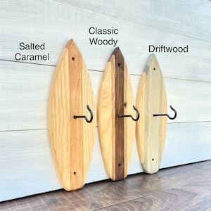 Peut inclure: Trois crochets muraux en bois en forme de planche de surf avec des crochets en métal. Les crochets sont teints dans différentes couleurs : "Salted Caramel", "Classic Woody" et "Driftwood".