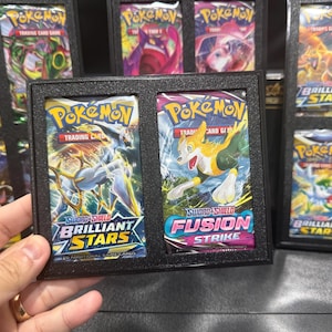 Può includere: Un espositore nero che contiene due buste di carte collezionabili Pokémon. Una busta è etichettata "Spada e Scudo Stelle Brillanti" e l'altra "Spada e Scudo Colpo Fusione". Altre buste sono visibili sullo sfondo.