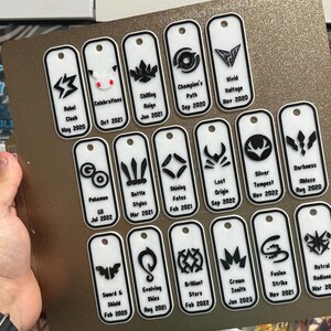 Könnte beinhalten: Eine Sammlung weißer rechteckiger Anhänger mit schwarzen Grafiken und Text, gehalten vor einem goldenen Hintergrund. Jeder Anhänger zeigt ein anderes Design sowie den Namen und das Datum eines Pokémon-Sets. Die Anhänger werden in einem Raster angezeigt.