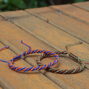 May include: Two braided bracelets. One is blue and orange, the other is green and brown. The bracelets are on a wooden surface. The bracelets are handmade and have a woven design.