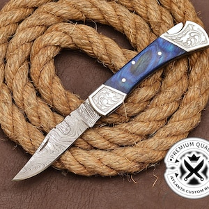 May include: A blue and silver pocket knife with a Damascus steel blade and a decorative handle. The knife is resting on a coil of rope. The knife has the text "Premium Quality Atlanta Custom Blades" on the side.